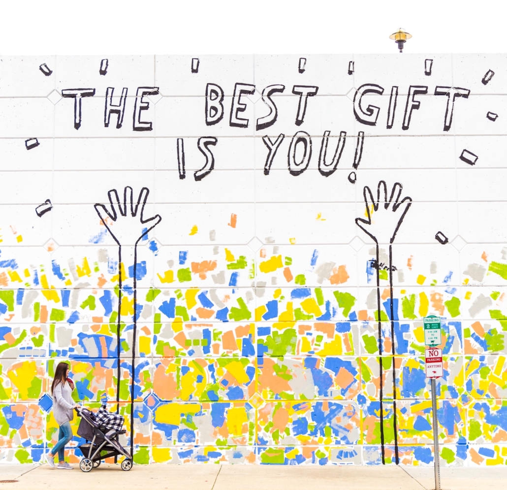 Woman pushing stroller past mural that says the best gift is you.